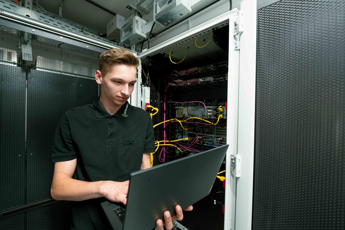 Foto aus Rechenzentrum zeigt Mann mit Laptop auf dem Arm vor einem geöffneten Serverschrank bei der Arbeit