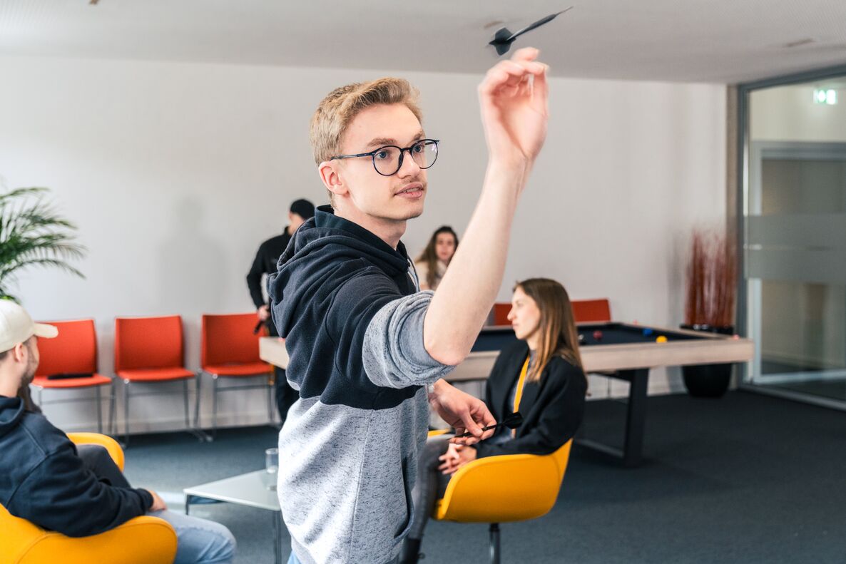 Gezeigt ist der Aufenthaltsraum der rockensteinAG, im Hintergrund steht ein Billardtisch, Mitarbeitende unterhalten sich, im Vordergrund wirft ein Mitarbeiter einen Dartpfeil auf eine Dartscheibe, welche sich hinter dem Fotografen befindet
