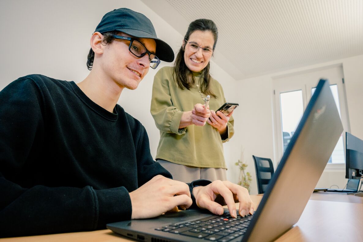 Foto aus unserem Coworking Space zeigt Mann arbeitend am Laptop und Frau hinter ihm mit freundlichem Blick und Lächeln