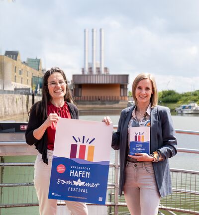Alexandra Rockenstein (rockenstein AG) und Sophia Hartl (Fachbereich Kultur Stadt Würzburg) auf der Pressekonferenz des Hafensommer 2025 halten ein Werbeplakat für 2025 vor des Kulisse des Hafensommers